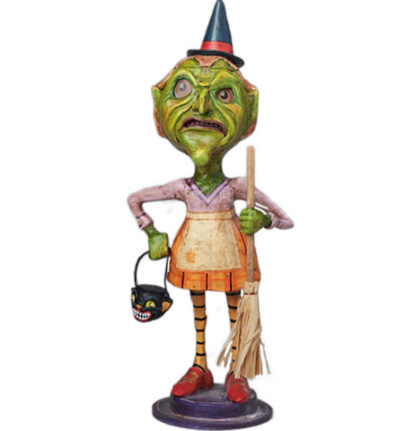 A figurine of a female green goblin wearing a black witches hat with red strap, pastel purple, yellow, and vibrant orange dress, yellow and black leggings, and red shoes. She is holding a candy bucket shaped liked a bats head and a broom.