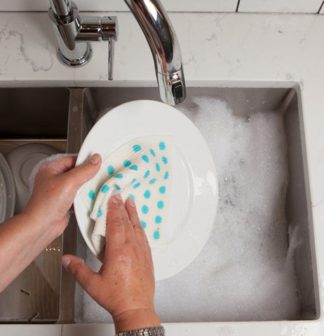 A sink full of dishes and soapy water, above which a hand uses a white cloth with blue spots to scrub a dish. 