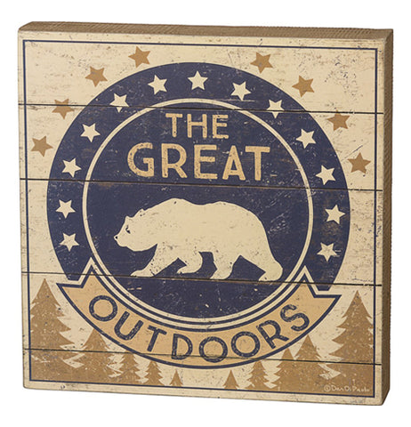 This small wooden box sign depicts a navy blue circle with a bear's outline. Around the circle are white and bronze colored stars against its beige background. The words, "The Great" are seen written in beige lettering while the word, "Outdoors" are written in black. Pine trees are shown in brown below the black and white bear symbol.