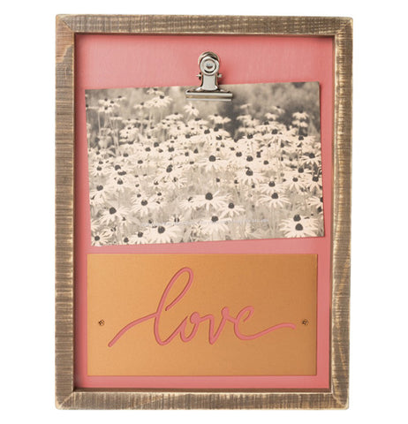 This is a wooden box shaped work of art on a peach pink wooden background inside the frame. There is a clip in the center holding onto a black and white picture of sunflowers and underneath the picture is a copper metal sheet attached with the word "Love" in cursive letters cut out.