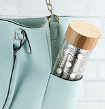 Glass travel mug with floral design with a tan bamboo lid and silver colored stainless steel infuser in a light blue purse in front of a white brick wall.