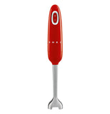 A handheld blender is stood on its' base. The body is red and the blade is stainless steel. It has an on/off button visible. The brand name "Smeg" on the body.