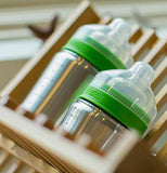 One 5 ounce and one 9 ounce brushed stainless baby bottle sitting on a wood dish drainer. Each bottle has a lid that is green where it screws to the bottle with a clear nipple and clear cover.