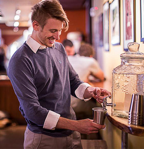 This image shows a young man in a blue sweater pouring water into the steel cup from a glass water dispenser.