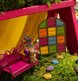 The multi-colored panel door is shown next to a pink bench in a small fairy garden setting.
