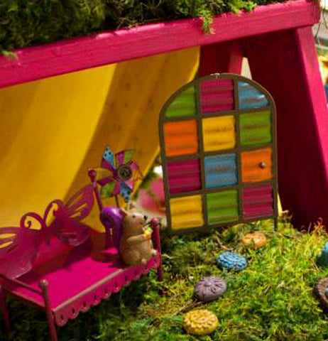 The multi-colored panel door is shown next to a pink bench in a small fairy garden setting.
