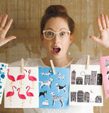 woman standing behind a clothesline with three Swedish dish cloths, one is white with Flamingos.,blue with dogs and white with grey buildings.