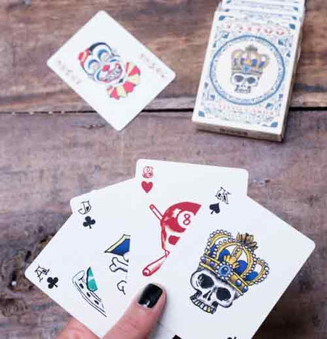 Hand holding King of spades card with skull wearing crown, Ace of hearts with red skull and crossbones, a jack of clubs, and an Ace of clubs are also in the hands over a brown wood table. The deck of cards with the king of clubs with skull and crown design next to the joker card with red, blue, and yellow circus clown design are on the table.