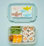 A transparent divided lunchbox lid has sharks on it. One shark is light brown, one is dark brown, one is yellow, and two are blue. Beneath it is the lunchbox itself, which is divided into three sections. On the left, the top section has green beans, and the bottom section has four orange slices and three small cookies. The right section has six pieces of sushi.