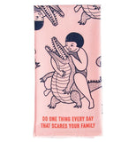This "Scares Your Family" dish towel features close-up image of a girl wrestling an alligator on pink background with a red text that says, "Do One Thing Every Day That Scares Your Family". 
