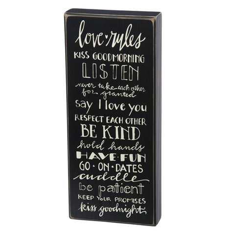 A rectangular black-painted wooden sign that has the words "Love Rules" in white at the top and all the rules listed below such as, Kiss Good Morning, Listen, Don't Take Each Other for Granted, Be Kind, Kiss Good Night, and more.