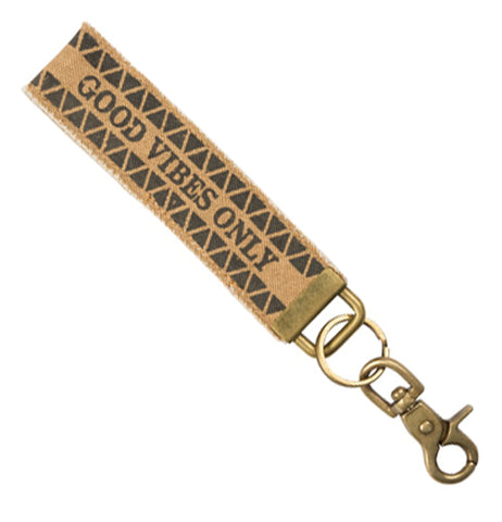 An 8" x 1" tan canvas key fob with the words "good vibes only" centered lengthwise and little black triangles in two rows above and beneath. Attached to the end is an antiqued brass D-ring with a brass key ring and antiqued brass lobster claw clasp hanging from it.