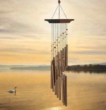 This garden ornament features 16 bronze tubes accented by stones hanging from a wooden hanger over a pond on a  sunset with a white duck swimming.