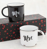 The black and white mugs are shown on a table; the black cup is on top of a black box with red flowers and the white cup is shown sitting below on the table next to the box.