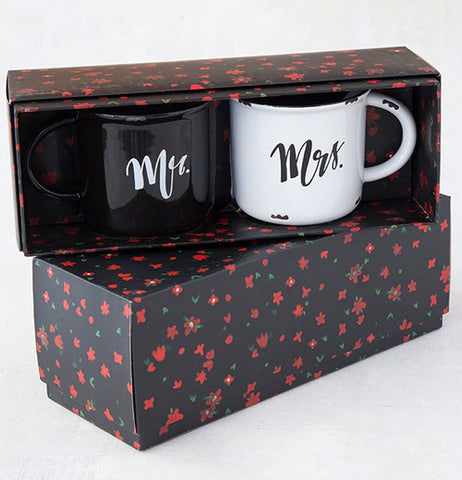 The black and white mugs are shown inside their packaging: a black box with red flowers. Another black box with red dots is shown sitting below the open box displaying the mugs.