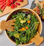 A round, light brown wooden salad bowl with wooden salad tongs. It's sitting on a counter with sald within it; a couple of serving trays, one with crackers and fruit and another with fruit on it, sit next to the bowl.