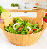 A light brown flared wooden salad bowl with wooden salad tongs. Its sitting on a counter filled with salad.
