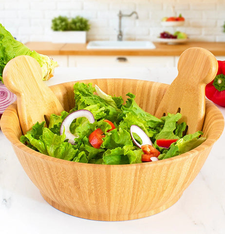 A light brown flared wooden salad bowl with wooden salad tongs. Its sitting on a counter filled with salad.