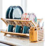 A wooden drying rack is being used. It has plates and coffee mugs in it. A wooden caddy has forks and knives in it.