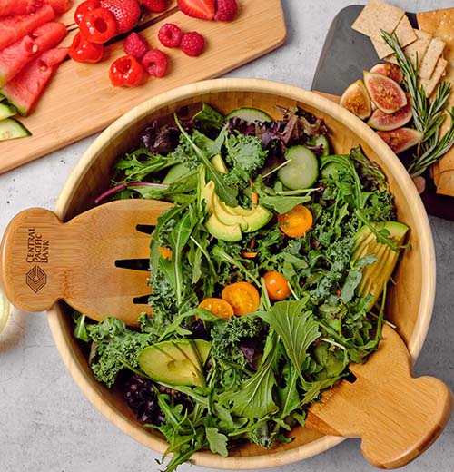 A wooden salad bowl with hand sized wooden salad tongs. It's sitting on a counter with sald within it; a couple of serving trays, one with crackers and fruit and another with fruit on it, sit next to the bowl.