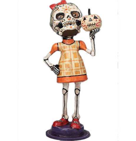 Marcella is a day of the dead inspired figurine holding a skull mask and holding a sugar skull pumpkin. wearing a yellow and orange plaid dress.