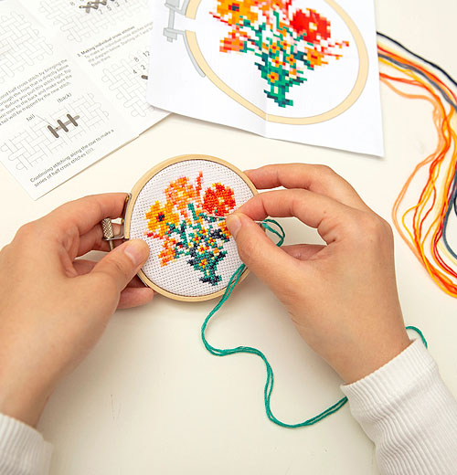 Someone embroidering a bouquet of flowers with a needle. The embroidery is attached to a small hoop and in the background you can see the pattern guide and the different colors of string used on the project.