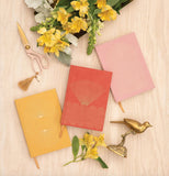 Three journals (one pink, one red, and one yellow) are arranged sylistically. Yellow flowers are arranged around the journals, along with pink and gold scissors and a gold bird statue.
