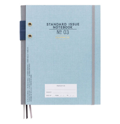 A light blue notebook has dark blue text ("standard issue notebook No. 03"), a gray elastic band to hold it closed, a gray spine with a blue elastic pencil holder, three bookmarks (two gray, one black), and a white "property of" sticker.