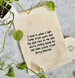 A beige tea towel with black text reading "I love it when I get home from work and my dog runs at me like we're finally going to nail that scene from Dirty Dancing." Laying on a marble counter with a vining plant at the top left and a wood coaster with a paw print under the bottom right corner of towel.