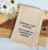 A beige dish towel with black text reading "Yesterday I quit drinking. But tonight we celebrate my comeback." laying on a wood board that is also laying on a marble counter. There is a clear cut glass vase with 2 white daisies in it.