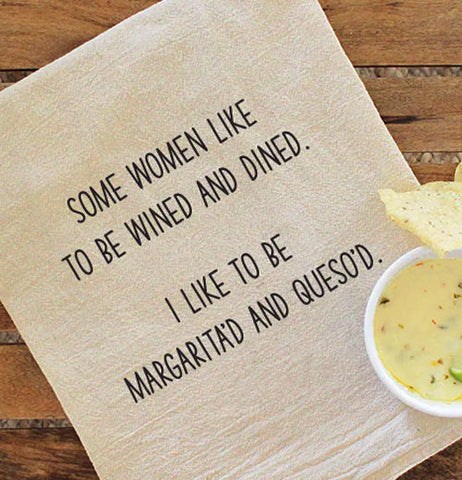 A beige dish towel with black text reading "Some women like to be wined and dined. I like to be margarita'd and queso'd." laying on a wood table with a bowl of dip and chips in the bottom corner.