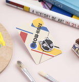 the pin is attached to a thick piece of paper close to the bottom right the paper has abstract shapes and lines with the words slanted along the middle left to the top right that say "Meow-haus" they are both sitting on a desk surrounded with art supplies like pencils, paint brushes, pallet and more.