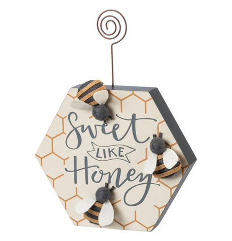 This hexagonal box sign has a yellow and white checkered pattern with cloth honey bee sculptures attached to it. In the middle of the box sign is gray text that reads, "Sweet Like Honey".