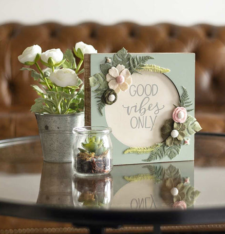 The green sign with the white circle and floral designs is shown sitting on a glass table next to some flowers.