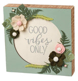 This green wooden box sign has a white circle in its middle surrounded by green and white flowers with green leaves. In the middle of the white circle are the words, "Good Vibes Only" in gray lettering. 