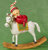 A young girl is riding a white rocking horse with a green halter and green and brown saddle pad. She wears a white, red, and green stocking cap, with red and white pajamas and socks. The background is green.