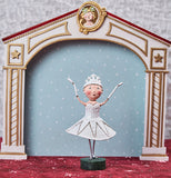 This Snow Queen is in a white dress with a tiara and holding her hands up on a stage.
