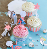 Three cupcakes made with the "Let's Be Mermaid" Cupcake Kit. Two cupcakes are topped with yellow frosting that has pink striped wrappers and one with the pink frosting that has the blue wave wrapper and also shows a mermaid, seashell and starfish cupcake toppers. 