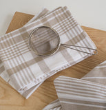 Three striped and plaid brown and white Sandstone Jumbo Dish Towels that are laying on a table with a metal flour sifter on top.