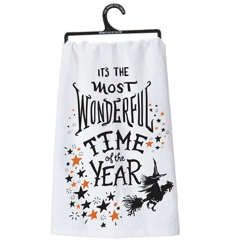 This white dish towel depicts the words, "It's The Most Wonderful Time of the Year" in black lettering. Below the letters is a black picture of a witch with a cat on a broomstick and surrounding the witch are black and orange pictures of stars.