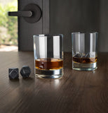 Two glasses of liquor are shown on a wooden table, one with two black ice cubes, the other having two black ice cubes outside it.
