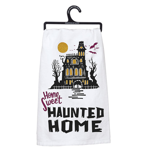 White dish towel with a black mansion that has glittery gold windows with a glittery gold moon and purple bats above. The words "Home sweet" are printed in purple and the words "Haunted Home" are printed in bold black letters underneath the mansion.