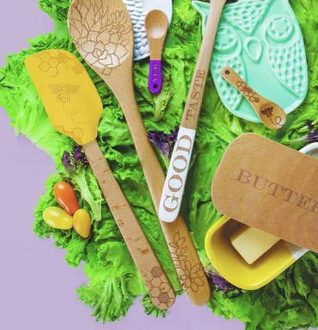 The yellow spatula with the bumblebee picture on its head is shown mixed with other spoons and spoon holders all sitting on grass.