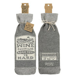 This cloth wine bottle cover is dark and light gray. On one side is a white sign with the words, "Wine Because Adulting Is Hard" in gray lettering and on the other side is a white sign with the words, "Warning: May Induce Joy" on the other. Below the joy sign is the logo "Primitives by Kathy".