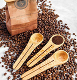 The coffee scoop with the picture of the owl is shown next to two other wooden spoons with similar designs. The three spoons sit on some coffee beans which sit next to a bag.