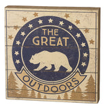 This small wooden box sign depicts a navy blue circle with a bear's outline. Around the circle are white and bronze colored stars against its beige background. The words, "The Great" are seen written in beige lettering while the word, "Outdoors" are written in black. Pine trees are shown in brown below the black and white bear symbol.