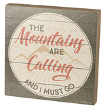 This small silver wooden box depicts the words, "The Mountains Are Calling And I Must Go" in its center with a cream colored and golden brown outlines of mountains behind the words. The words, "Mountains" and "Calling" are written in orange red lettering, while the rest of the sentence is in grey lettering.