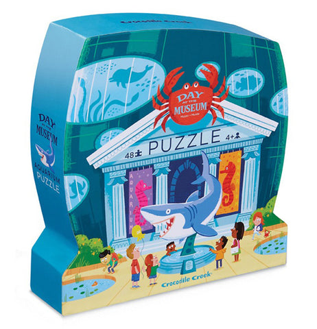 The "Aquarium Day at the Museum" Puzzle kit has 48 pieces in a blue box that is shaped like a museum. It has the image of a museum with sea creatures displayed and children looking at the dispalys. 