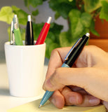 A person's hand is shown writing on some paper with one of the pens. Three more pens are seen inside a cup.