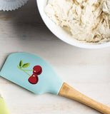 The turquoise spatula with the design of the two connected cherries is shown lying on a table next to a bowl of batter.